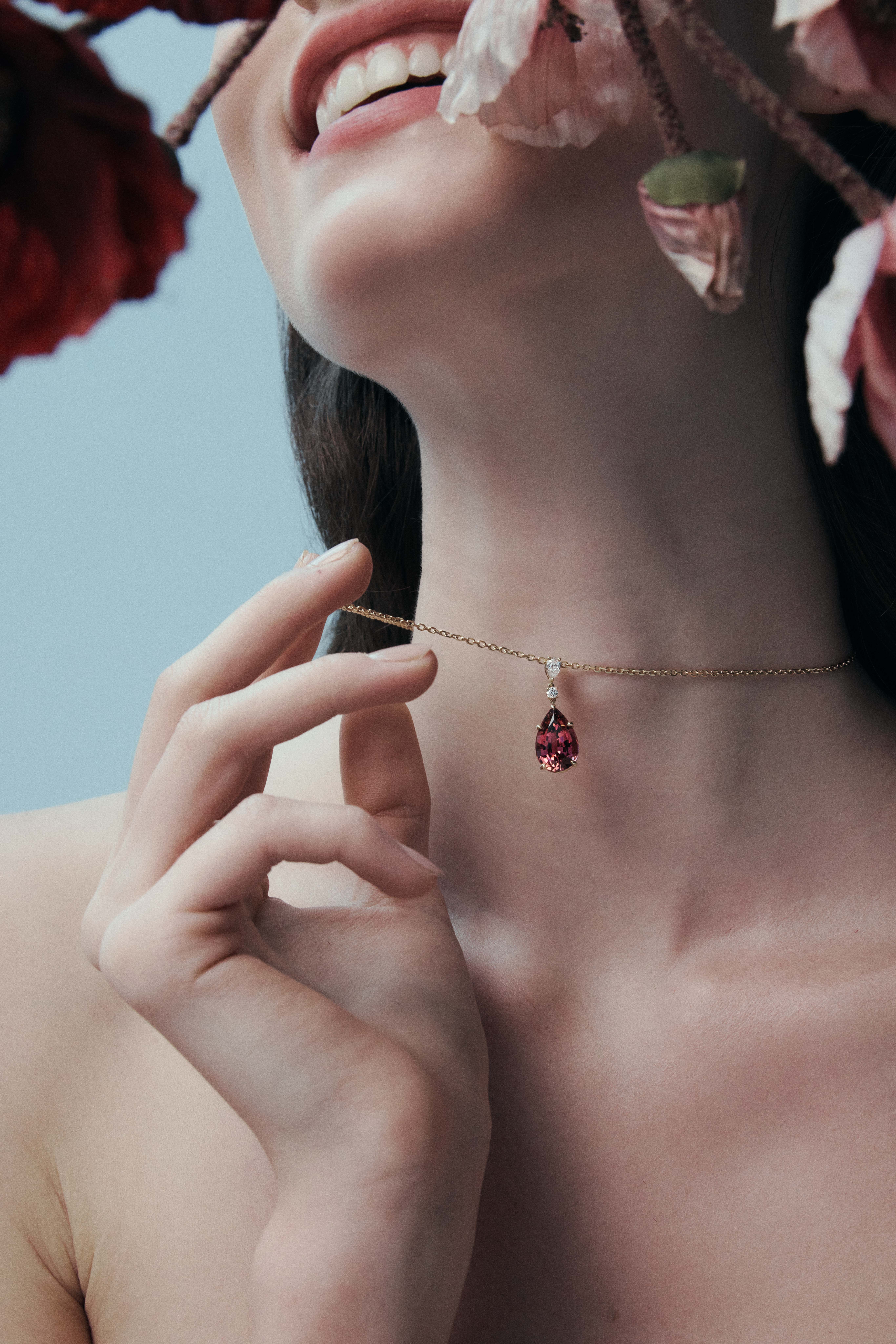 A woman with fair skin wears a gold chain necklace with a pink gemstone, accessorized with dark pink flowers against a blue background.