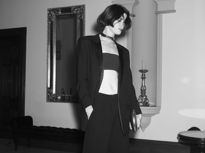 A monochrome photograph of a woman in a black blazer and pants with hands in pockets, standing indoors with a mirror and decorative column.
