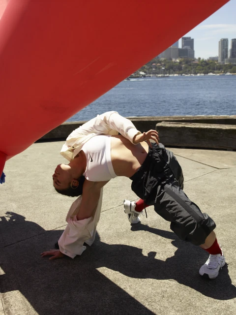 Adult female in a white sports bra and dark bottoms does a yoga pose outdoors with a red structure against a city and ocean backdrop.