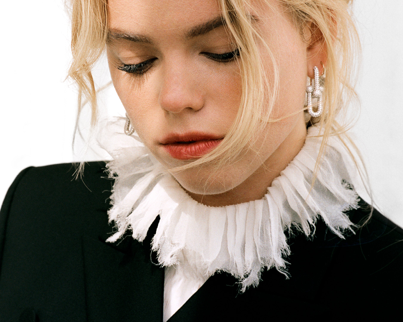 Close-up portrait of a woman with blonde hair wearing a white ruffled collar and black jacket with diamond hoop earrings