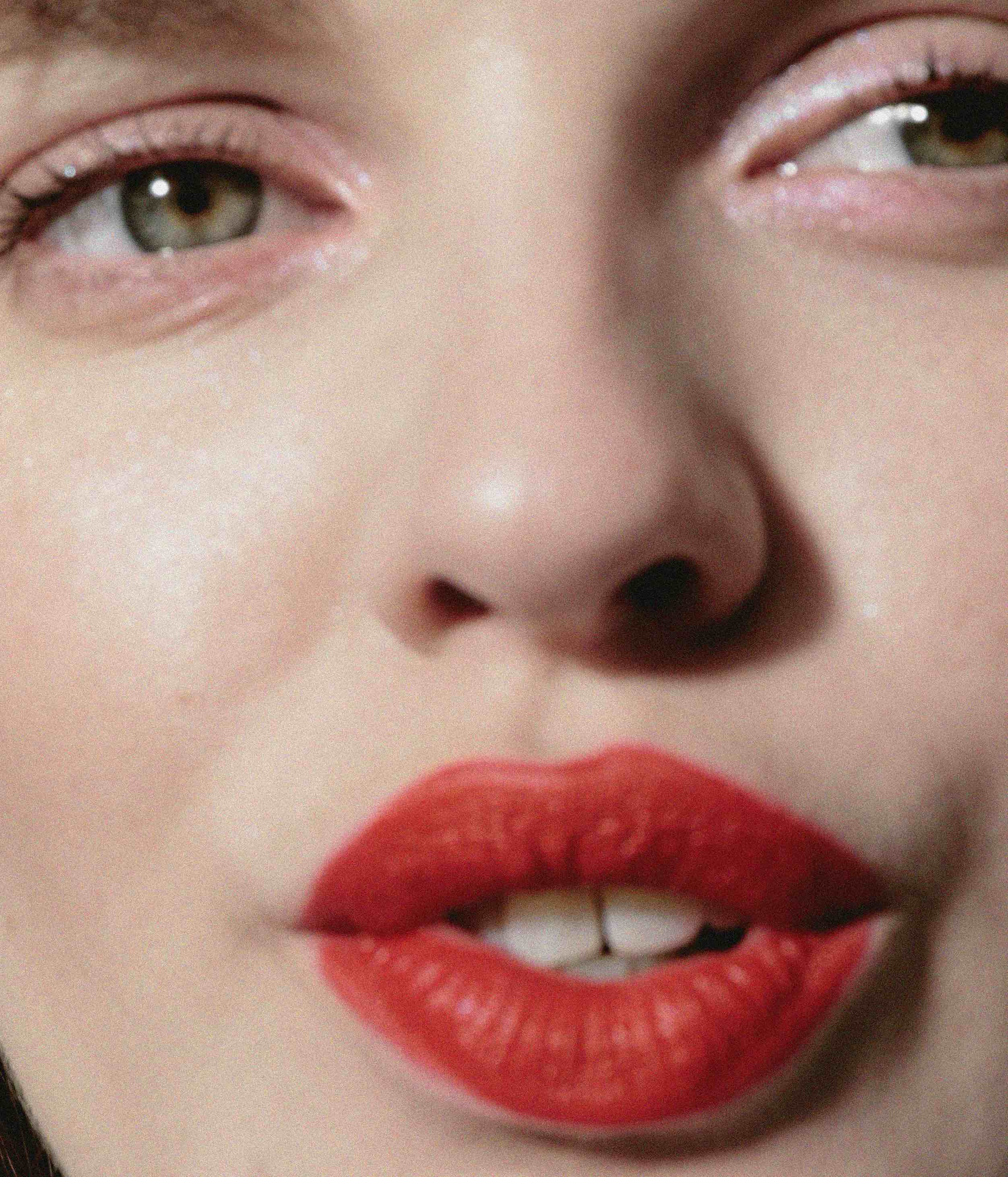 Close-up of a young adult female's face featuring red lipstick, visible teeth, and olive-green eyes in a beauty-oriented portrait.