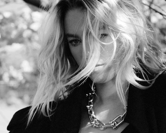 Close-up monochrome portrait of a woman wearing a heavy metallic chain necklace and dark jacket with blonde hair blowing across her face