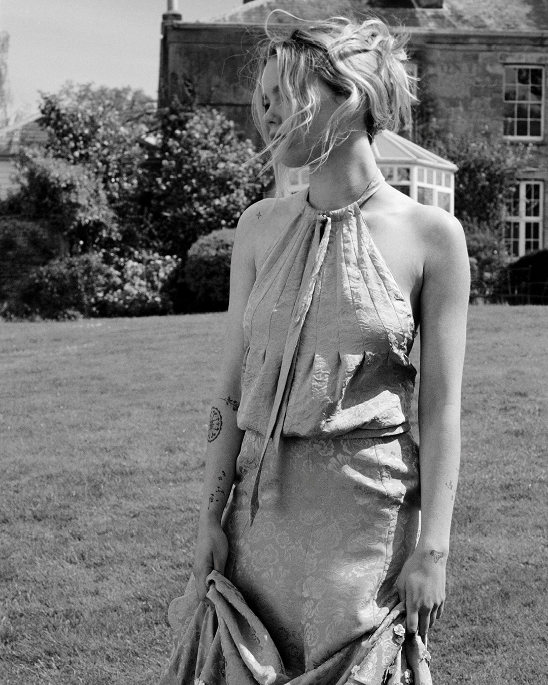 Monochrome portrait of a woman with an updo hairstyle wearing a patterned halter dress on a lawn outside a stone estate.