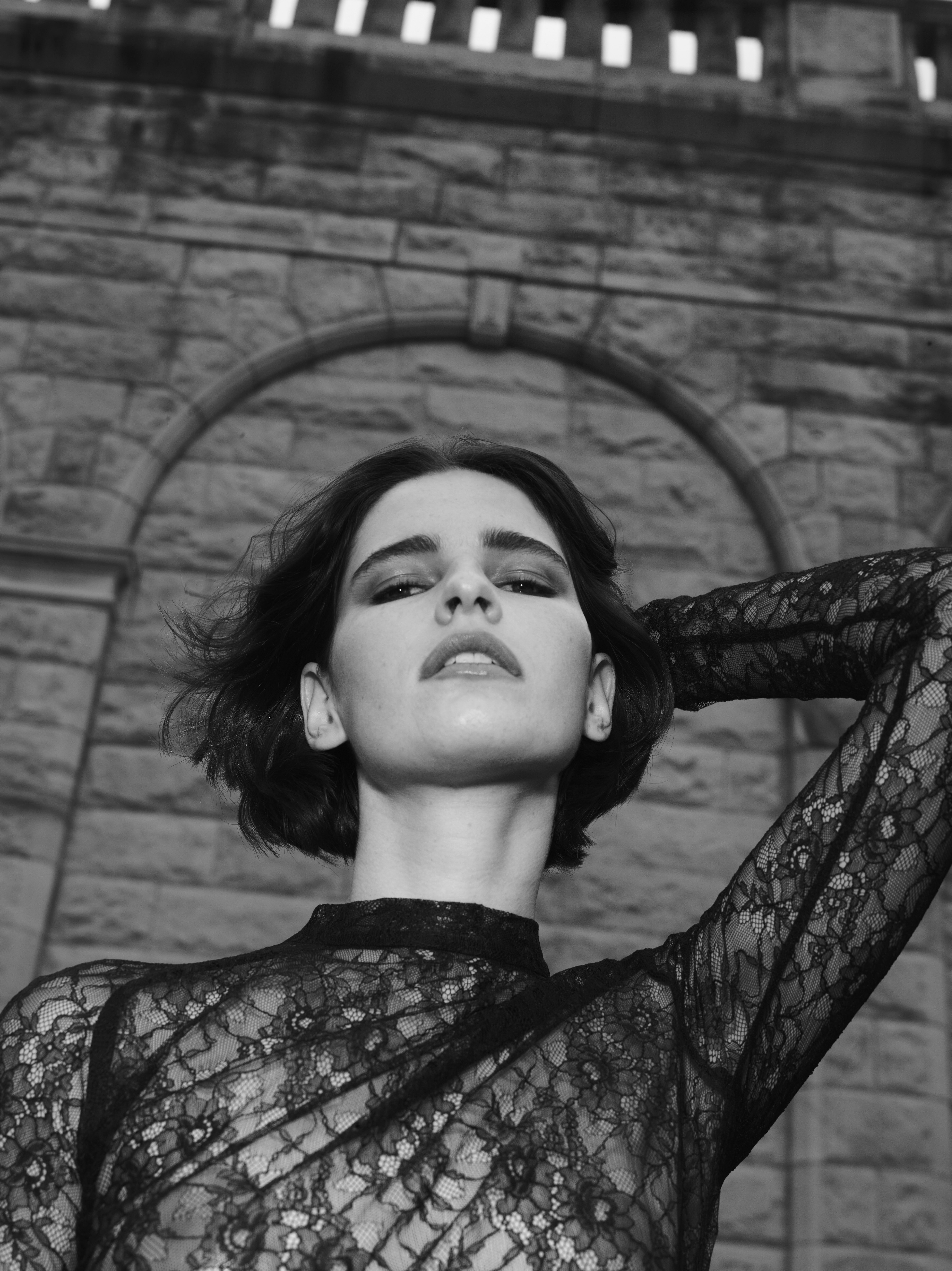 Black and white portrait of a woman in lace top standing in front of a stone archway, looking up at the camera with neutral expression.