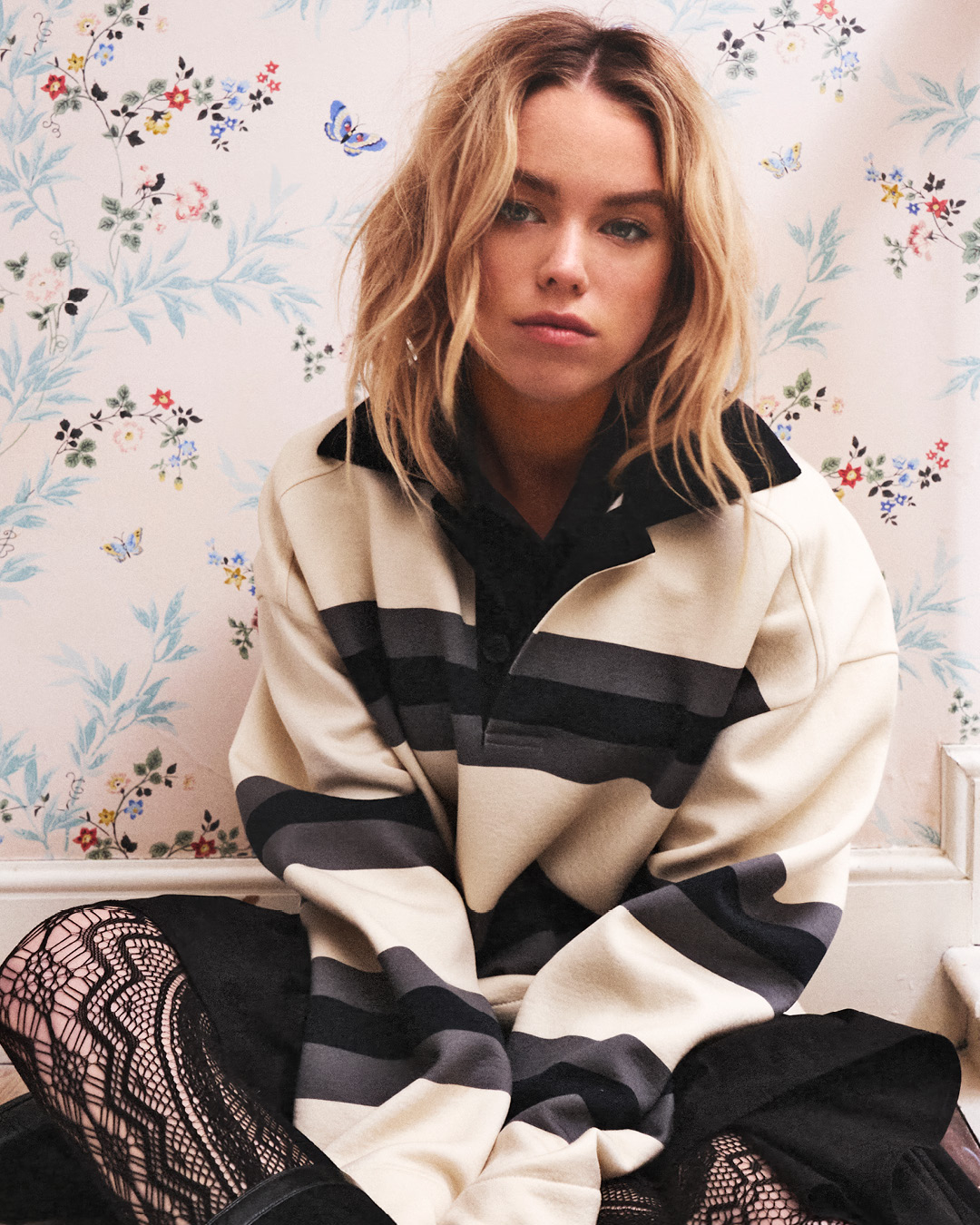 Woman with wavy blonde hair sits indoors wearing a striped long sleeve and black skirt in front of floral wallpaper. Fashion shoot.