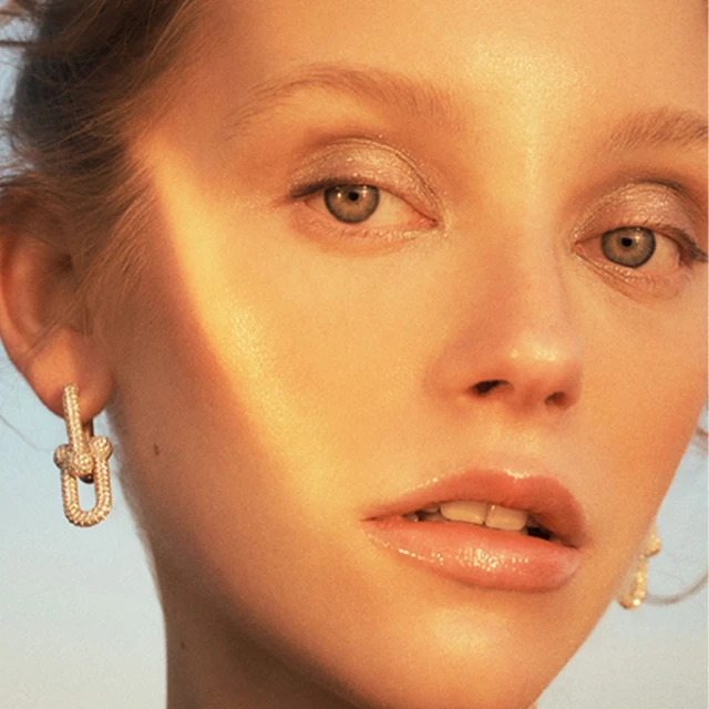 Close-up portrait of a woman with fair skin, gold eyeshadow, and diamond earrings. The golden light enhances her facial features.