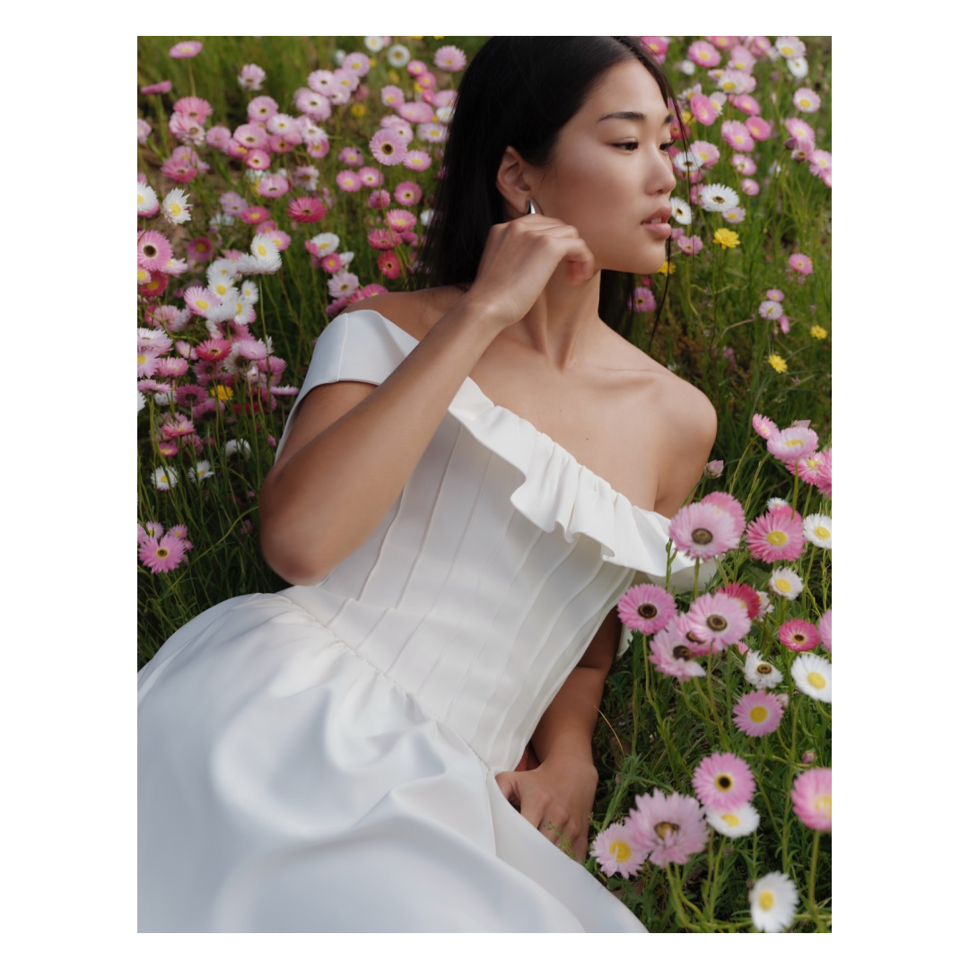 A woman in a white off-the-shoulder dress is lying in a field of colorful daisies. Soft light illuminates the scene.
