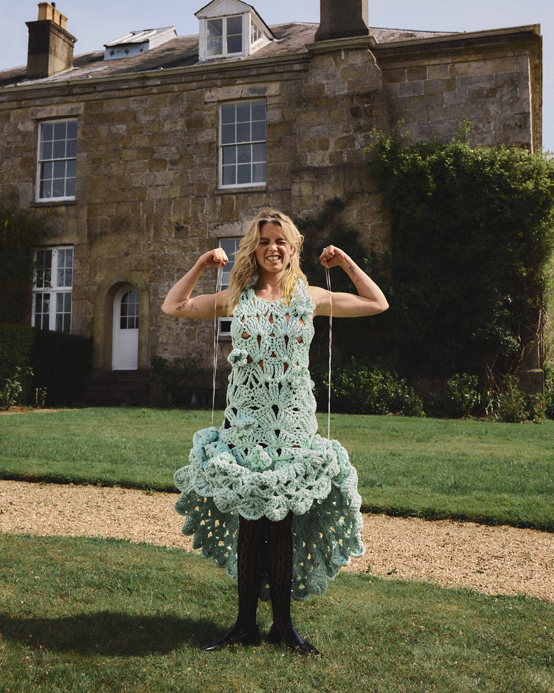 A smiling adult woman poses outdoors in a crochet dress against a backdrop of a large stone house and green lawn, flexing her biceps.