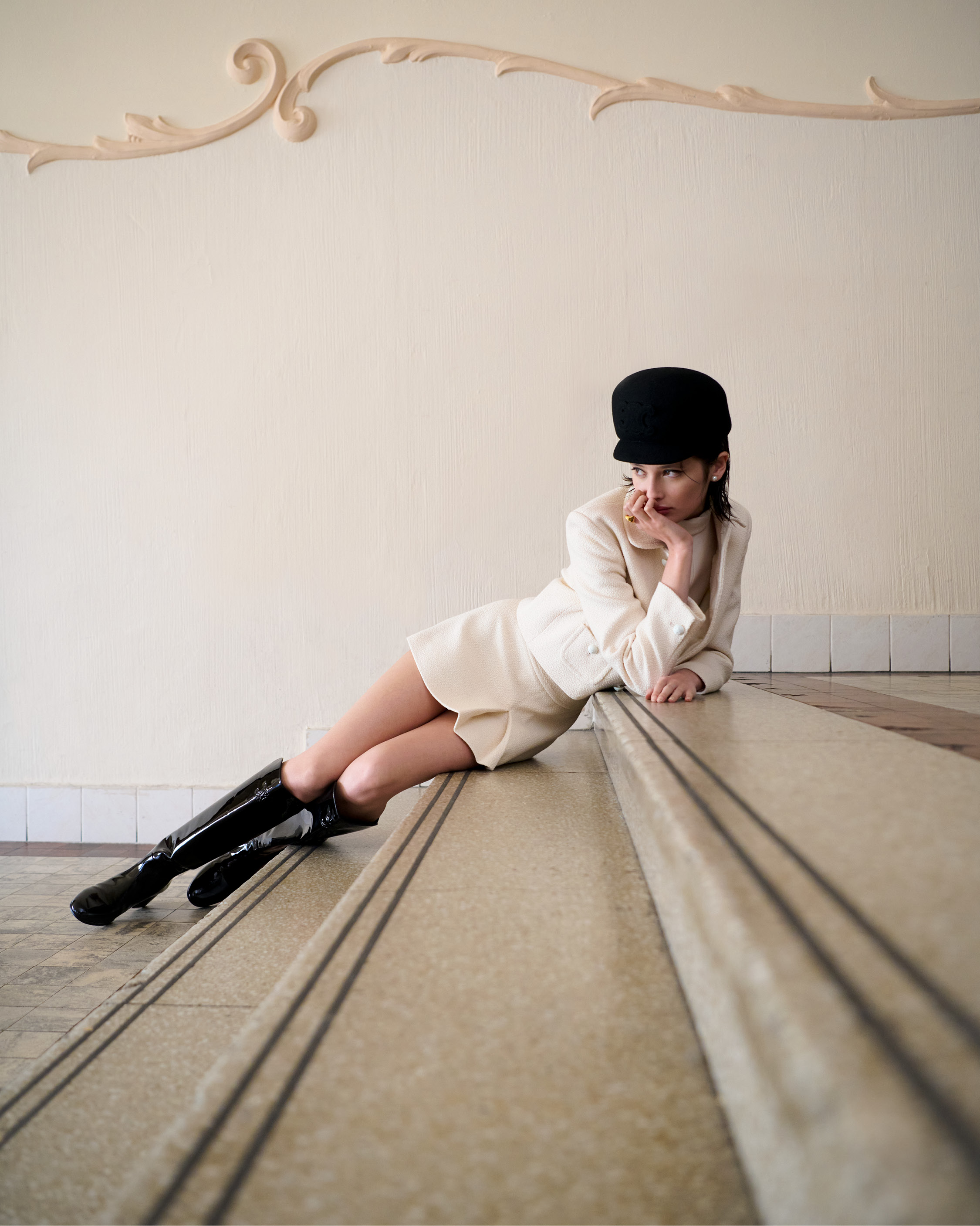Woman in a white dress and black boots poses on marble stairs, complemented by ornate wall detail and natural light.