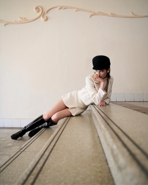 Woman in a white dress and black boots poses on marble stairs, complemented by ornate wall detail and natural light.