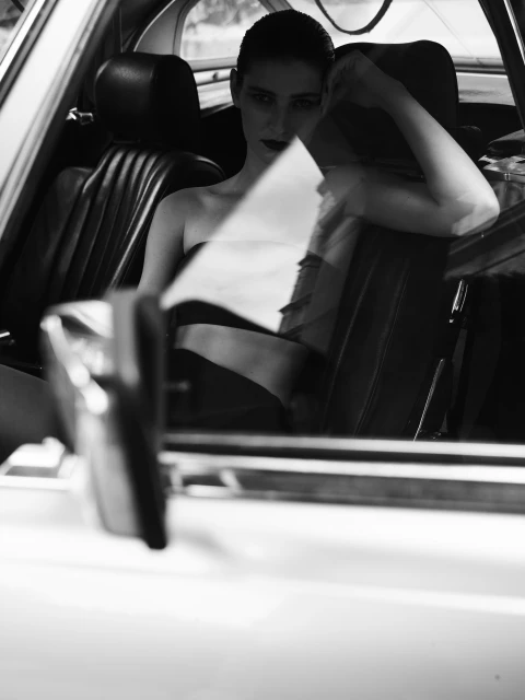 Monochrome portrait of a woman with dark makeup in a vintage car interior, reflected in a car window, capturing a timeless fashion aesthetic.