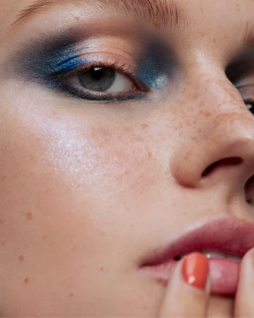 Extreme close-up of a model's face with dramatic blue eye makeup, visible freckles, and glossy lips.