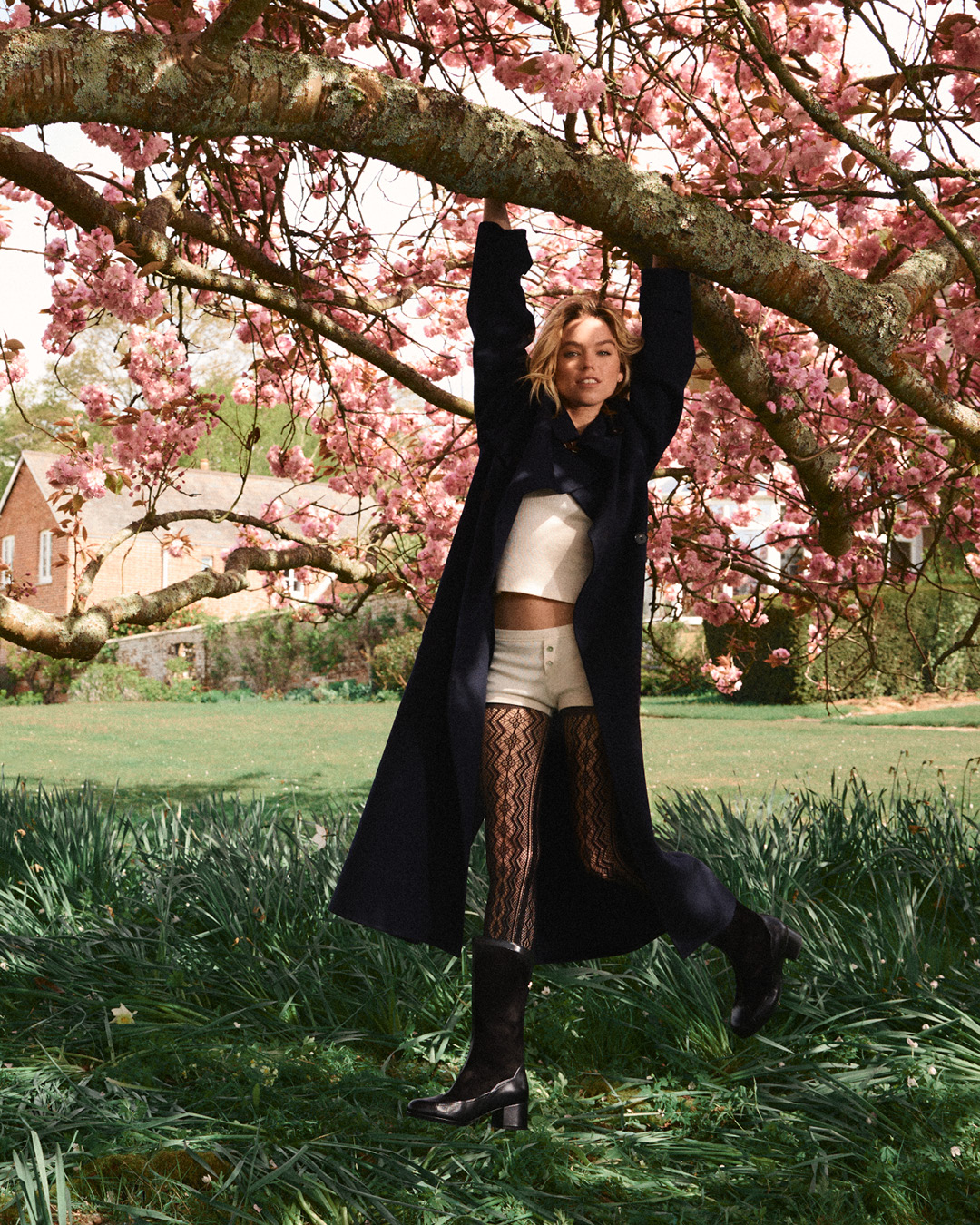 A woman hangs from a cherry blossom tree branch wearing a long dark coat, corset top, shorts, patterned tights, and platform boots.