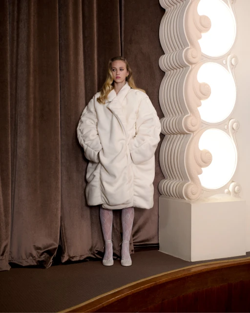 A woman in a white coat stands against a brown curtain and ornate white column in a fashion portrait, with focus on her coat and outfit.