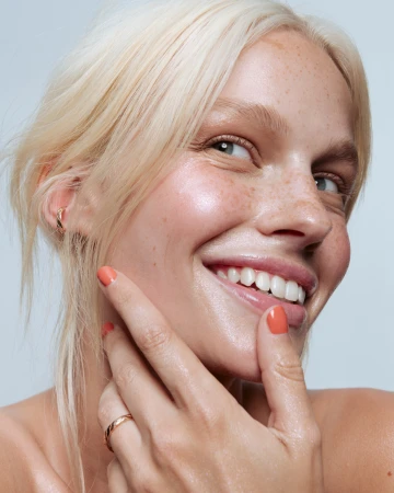 Close-up portrait of a smiling blonde woman with freckles and glossy coral nail polish touching her face