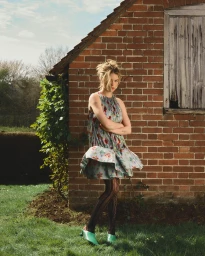 Woman in floral print mini dress and patterned tights standing outdoors against a red brick wall