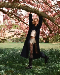 Woman hanging from a blooming cherry blossom tree branch in a garden, wearing a long dark coat, white knit shorts, and boots.