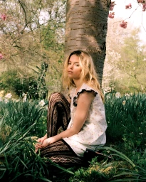 Young woman in a patterned white dress and black patterned tights kneeling at the base of a large tree in a spring meadow.