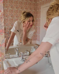 Young woman leaning over a bathroom sink looking into a mirror, wearing a white collared shirt.