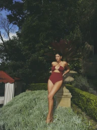 Woman in a burgundy one-piece swimsuit leaning against a stone planter in a lush garden by Eminente Creative Production