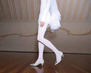 Close-up of a person in white tights and white heels walking past an ornate wall molding.