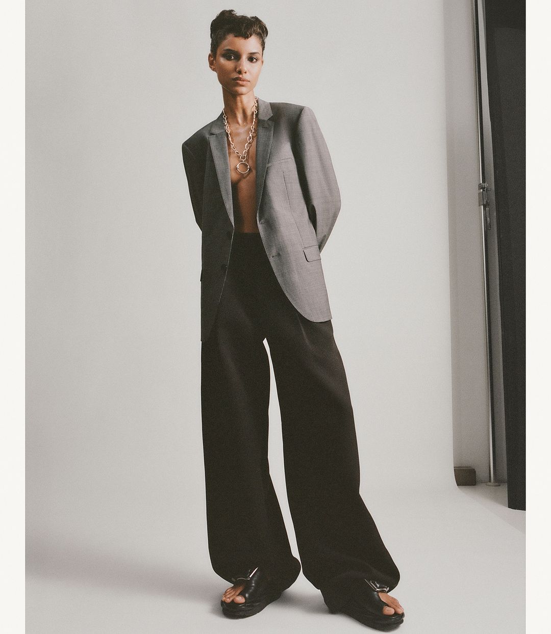 Fashion shot of a slender woman in an oversized grey blazer, wide-leg black trousers, and sandals, standing in a bright studio.