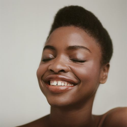 Close-up shot of a smiling African-American woman with closed eyes, radiant skin, and a natural hairstyle against a plain backdrop.