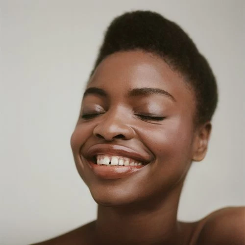 Close-up shot of a smiling African-American woman with closed eyes, radiant skin, and a natural hairstyle against a plain backdrop.