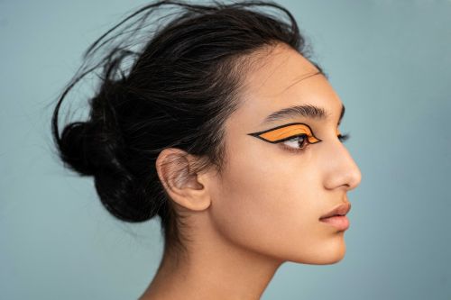Profile portrait of a woman with dark hair and bright orange graphic eye makeup, against a light blue background in a beauty shot.