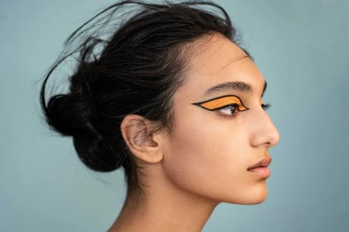 Profile portrait of a woman with dark hair and bright orange graphic eye makeup, against a light blue background in a beauty shot.