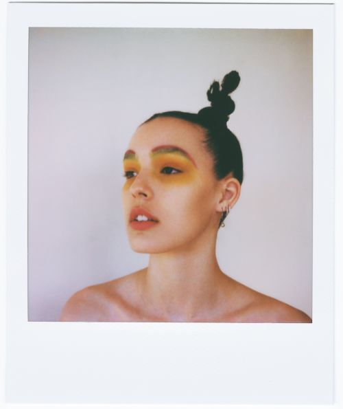 Portrait of a young adult female with bright yellow eyeshadow and hair styled in a topknot, posing in a light, neutral indoor setting.