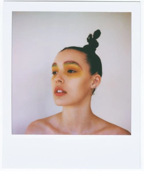 Portrait of a young adult female with bright yellow eyeshadow and hair styled in a topknot, posing in a light, neutral indoor setting.