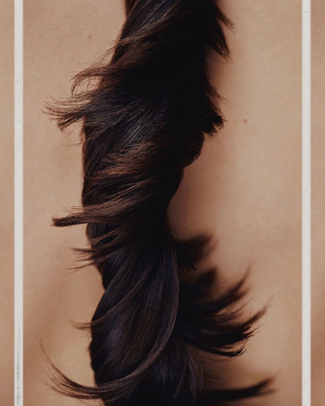 Close-up of long, dark hair twisted and styled against a light beige background with white vertical lines on the edges of the frame.