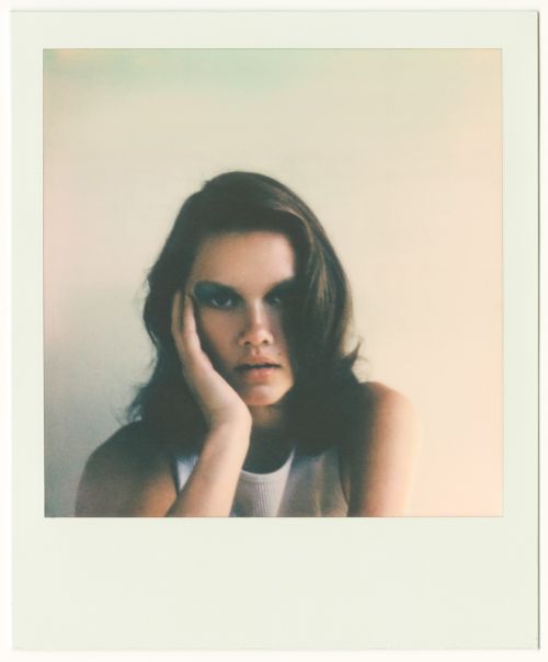 A young woman with dark hair, wearing a white tank top, poses in a Polaroid-style photo with an off-white background and a dreamy aesthetic.