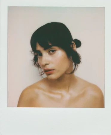 Portrait of a woman with dark hair pulled back, facing the camera in a close-up studio shot, with neutral background and soft lighting.