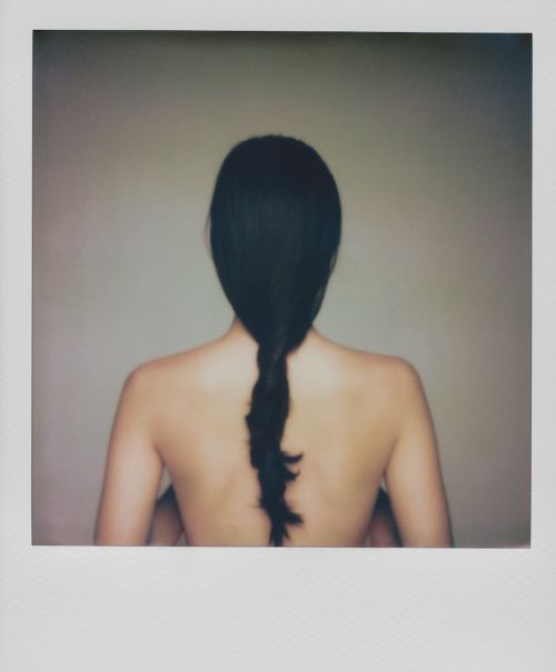 Back view of a woman with dark hair braided down her back. Neutral background with soft lighting accentuates the subject.