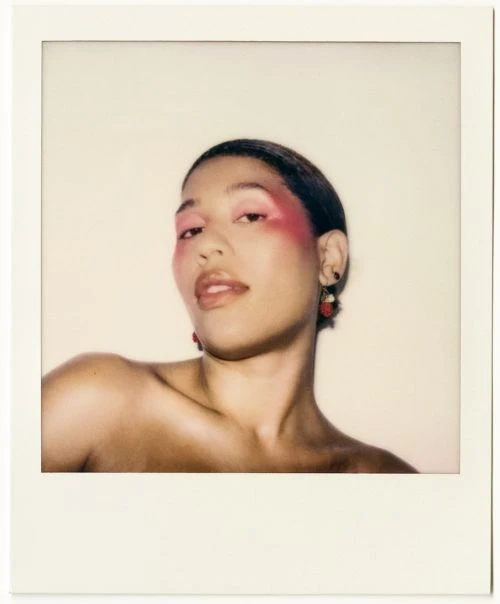 Portrait of a woman with pink eyeshadow and red drop earrings, framed in a white Polaroid border against an off-white background.