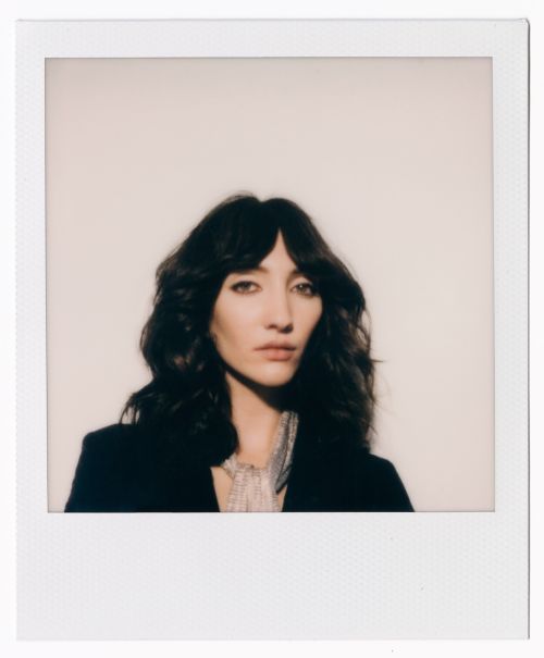Close-up of a brunette woman with bangs wearing a black jacket against a neutral background in a polaroid frame, fashion portrait.