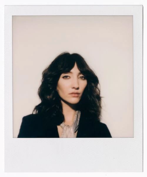 Close-up of a brunette woman with bangs wearing a black jacket against a neutral background in a polaroid frame, fashion portrait.