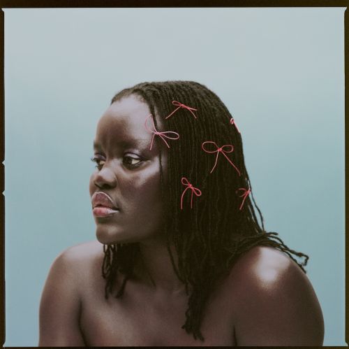 Close-up portrait of an adult Black woman against a blue background, adorned with small pink bows in her dark hair, looking off-camera.
