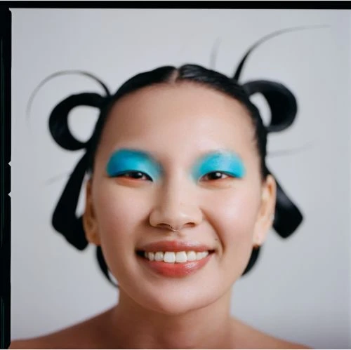 Close-up of an Asian woman with a wide smile, bright blue eyeshadow, and an elaborate hairstyle with black ribbons.