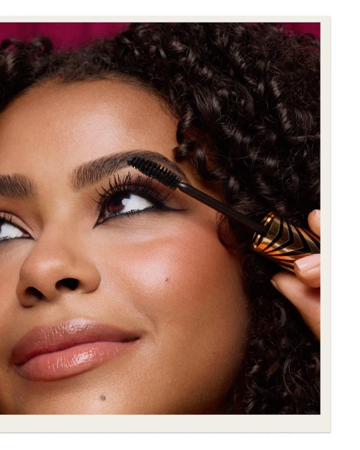 Close-up of a woman with brown curly hair applying mascara. Beauty shot in a studio with a dark-pink background.