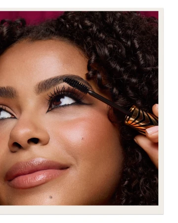 Close-up of a woman with brown curly hair applying mascara. Beauty shot in a studio with a dark-pink background.