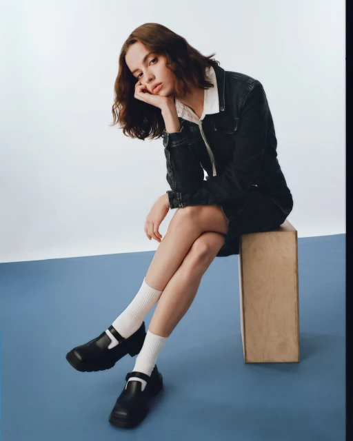 A young woman with brunette hair poses indoors. She is sitting on a wooden block wearing a denim jacket and Mary Jane shoes.