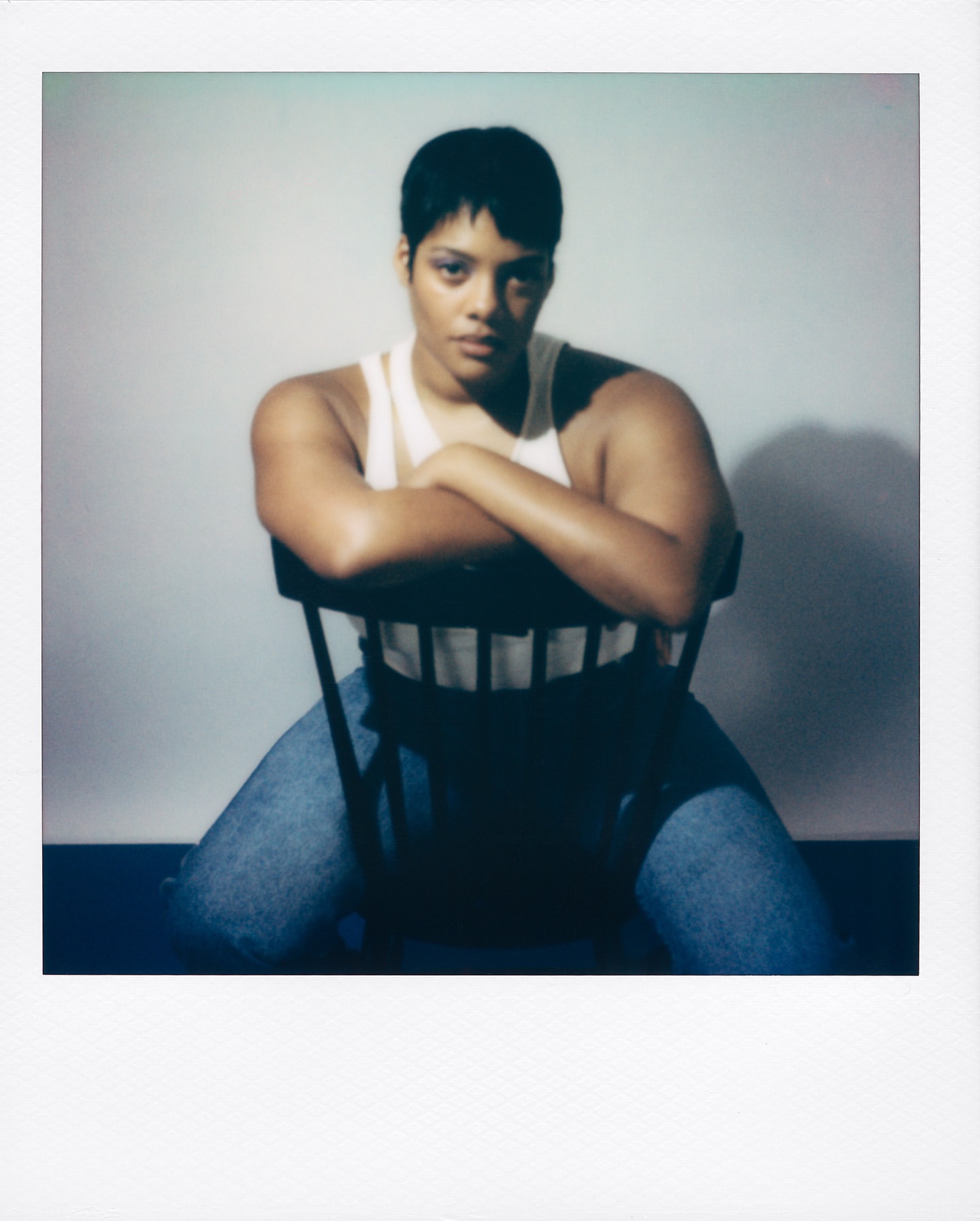 Portrait of an adult female with short dark hair, sitting in a chair in a photo studio, wearing a white tank top and blue jeans.