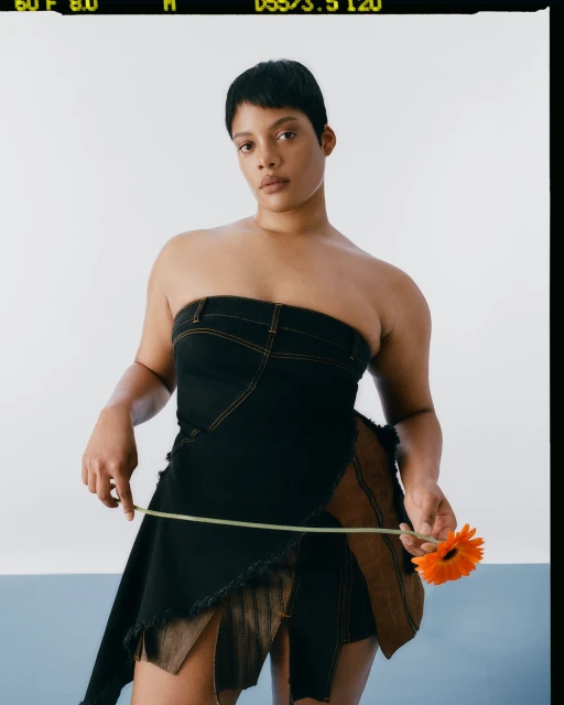 A short-haired woman in a strapless dress holds an orange gerbera daisy in a studio shot against a color-blocked white and blue background.