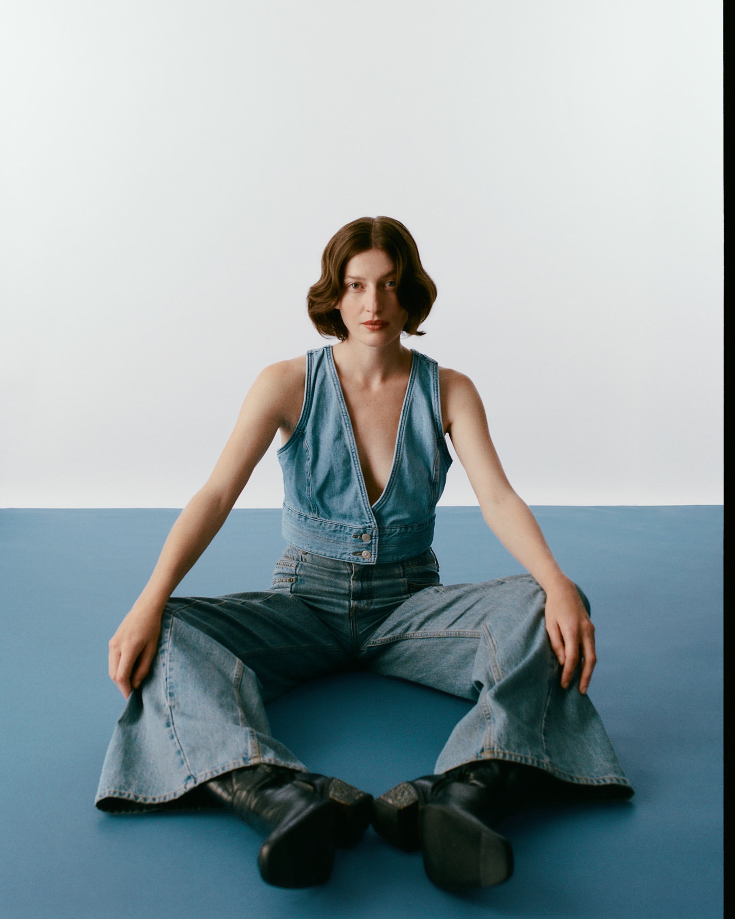 A young woman with bobbed hair sits on a blue surface wearing a denim vest, matching jeans, and black boots against a white backdrop.