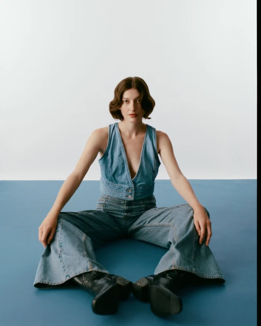 A young woman with bobbed hair sits on a blue surface wearing a denim vest, matching jeans, and black boots against a white backdrop.