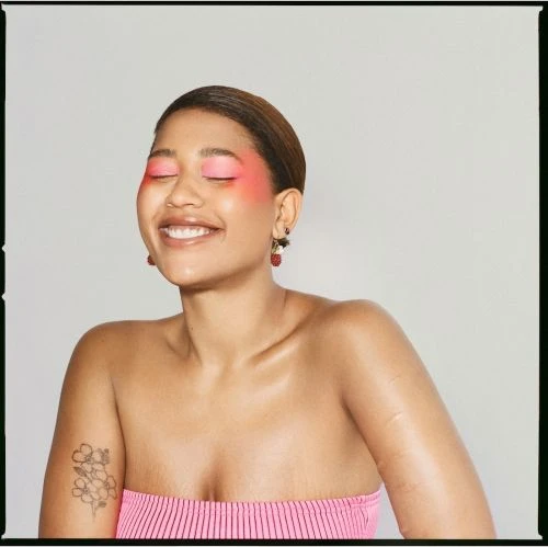 Close-up studio portrait of a smiling woman with red eye makeup and pink top, looking upward with a tattoo on her arm.