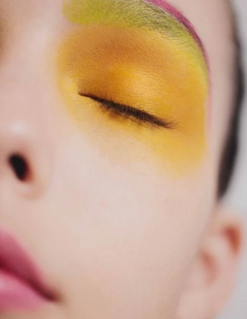 Close-up of an adult female face with vibrant yellow eyeshadow extending to the brow bone, accented with a hint of pink eyebrow makeup.